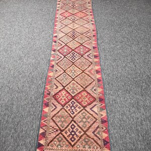 May include: A long, narrow runner rug featuring a repeating diamond pattern in shades of pink, red, brown, and blue. The rug has a geometric border with triangular motifs and a fringed edge at the bottom, showcasing a textured, woven construction.