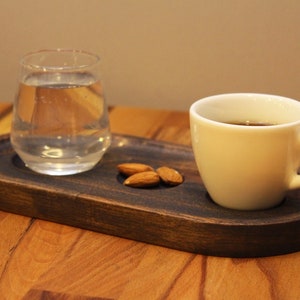 以下が含まれることがあります： 楕円形の木製サービングトレイには、水を入れたグラス、小さな白いコーヒーカップ、3つのアーモンドが置かれています。 コーヒーカップには濃い液体が入っています。