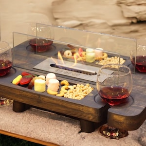 Puede incluir: Una bandeja de madera teñida oscura con una chimenea incorporada, que contiene cuatro copas de vino tinto, aperitivos y un protector contra el viento de vidrio. La bandeja tiene huecos para las copas de vino y está colocada sobre una mesa baja.