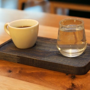 May include: A white ceramic coffee cup filled with dark liquid and a clear glass of water sit on a dark wooden tray. The tray is rectangular with a recessed area for the cup and glass. The background is a wooden table.