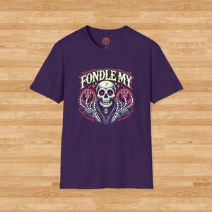 May include: Purple t-shirt with a graphic of a skeleton holding two glowing orbs. The text "FONDLE MY" is above the skeleton.