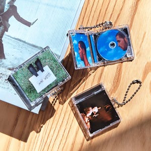 Könnte beinhalten: Miniatur-CD-Hüllen aus klarem Kunststoff mit verschiedenen Albumcovern und einer Kette. Ein Case zeigt eine blaue CD und Albumcover mit einem Männerporträt. Ein anderes hat ein kleines Grasstück mit schwarzen Stiefeln und einem Zettel mit der Aufschrift "WATER BOOTS".