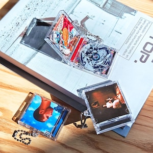 May include: Clear acrylic CD case keychains with various album art inserts. One keychain features a cityscape design, another a portrait against a blue sky, and a third with a sepia-toned image. Each keychain has a silver ball chain.