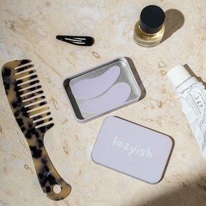 Op de afbeelding: Een assortiment schoonheids- en zelfzorgartikelen, waaronder een schildpadkam, een zwarte haarclip, een kleine parfumfles, een tube crème en een blikje oogpatches. Een tweede blikje is voorzien van het label "lazyish". De items zijn gerangschikt op een lichtgekleurd oppervlak.