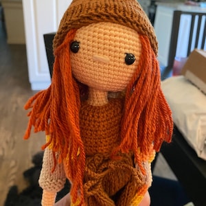 Könnte beinhalten: Handgemachte Häkelpuppe mit orangefarbenem Garnhaar, schwarzen Knopfaugen und einer kleinen Nase. Die Puppe trägt einen braunen Hut, ein braunes Kleid und einen cremefarbenen Pullover mit gelben und orangefarbenen Akzenten. Ein einzigartiger, handgefertigter Artikel.