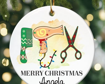 May include: White ceramic Christmas ornament with a watercolor background. The ornament features a green comb, a pink hair dryer, and red and black scissors. The text "Merry Christmas Angela" is printed on the ornament.