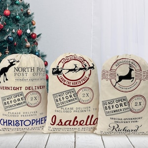 May include: Three beige canvas Santa sacks with red and black printed designs. The sacks are labeled "North Pole Post Office", "Express Delivery from the North Pole", and "Reindeer Mail". Each sack has a personalized name tag: "Christopher", "Isabella", and "Richard".