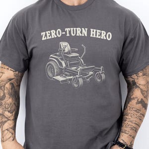 Op de afbeelding: Donkergrijs t-shirt met de tekst "ZERO-TURN HERO" boven een afbeelding van een zitmaaier. Het ontwerp is in een lichtbeige kleur. Het shirt heeft een ronde hals en korte mouwen.