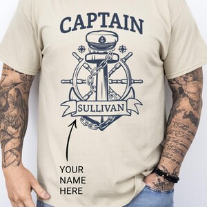 Sand Colored cotton t-shirt with bold navy graphic featuring a captain’s hat, anchor, ship wheel, and personalized name banner reading “SULLIVAN,” worn by a tattooed man; custom captain shirt ideal for boat owners or as a Father’s Day gift.