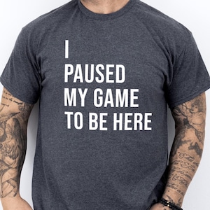 May include: A dark heather grey t-shirt with the text "I paused my game to be here" printed in white.