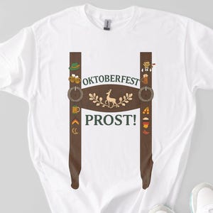 White cotton t-shirt with a printed lederhosen suspender design featuring festive Oktoberfest icons like beer mugs, pretzels, sausages, and a central deer graphic. The shirt displays the words "Oktoberfest" and "Prost!" in green text on the chest.