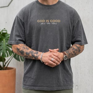 May include: A charcoal grey short-sleeve t-shirt with a crew neckline. The chest features the text "GOD IS GOOD" in a bold, light-coloured sans-serif font, with the words "All The Time" written underneath in a smaller, cursive script font.