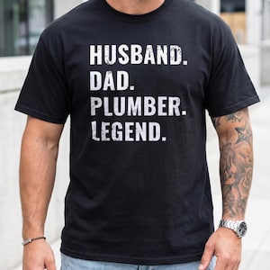 May include: Black t-shirt with white text: HUSBAND. DAD. PLUMBER. LEGEND. The short-sleeved shirt is likely cotton. The person wearing the shirt has a beard and a watch.