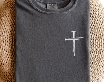 May include: A charcoal gray t-shirt with a white embroidered cross design on the front. The shirt is folded neatly and rests on a woven, circular surface. The t-shirt has a crew neck and short sleeves.