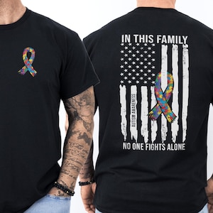 May include: Black t-shirt featuring a colorful puzzle ribbon, a symbol of autism awareness. The back of the shirt displays an American flag design with the text "IN THIS FAMILY, AUTISM AWARENESS, NO ONE FIGHTS ALONE."