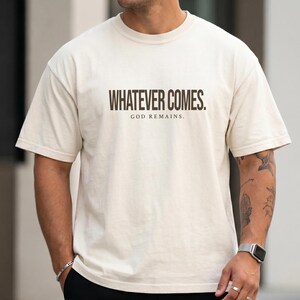 Może przedstawiać: Kremowy t-shirt z krótkim rękawem, na którym znajduje się ciemnobrązowy napis "WHATEVER COMES." pogrubioną czcionką bezszeryfową, a mniejszy napis "GOD REMAINS." wydrukowano bezpośrednio pod nim w pasującym ciemnobrązowym kolorze.