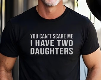 Może przedstawiać: Czarny t-shirt z białym napisem "YOU CAN'T SCARE ME I HAVE TWO DAUGHTERS". Koszulka z krótkim rękawem ma okrągły dekolt. Osoba nosząca koszulkę uśmiecha się.