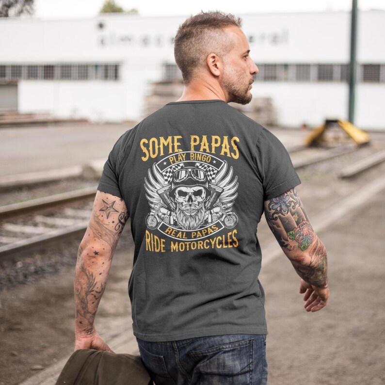 Puede incluir: Una camiseta gris oscuro con un dise&ntilde;o gr&aacute;fico de una calavera con barba y alas. El texto en la camiseta dice "Some Papas Play Bingo Real Papas Ride Motorcycles".