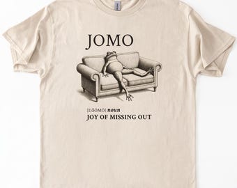 Może przedstawiać: Beżowa koszulka z napisem "JOMO" nad ilustracją żaby na kanapie. Poniżej tekst "[dʒoʊmoʊ] rzeczownik RADOŚĆ Z OMIJANIA".