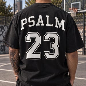 May include: A black short-sleeved t-shirt featuring the word PSALM in white capital letters arched across the upper back, with the number 23 printed in large white block numerals with a black outline centred below the text on the back.