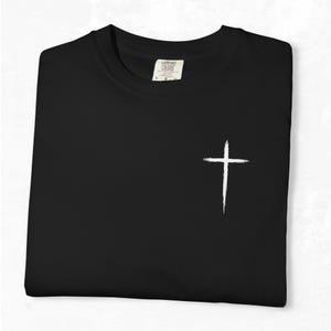May include: Black t-shirt with a white cross graphic on the front. The shirt is folded neatly, and the tag reads "Comfort Colors". The cross is a simple, brushstroke design.
