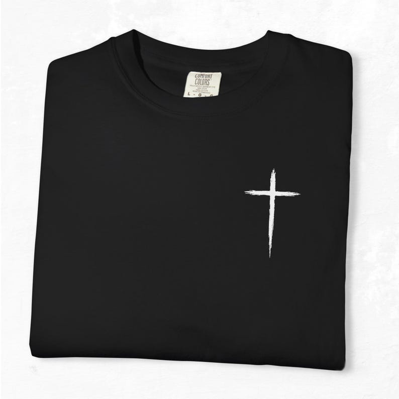 May include: Black t-shirt with a white cross graphic on the front. The shirt is folded neatly, and the tag reads "Comfort Colors". The cross is a simple, brushstroke design.