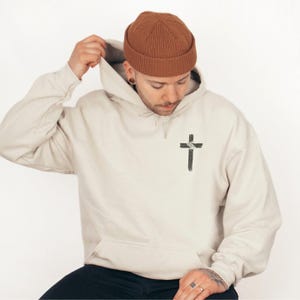 K&ouml;nnte beinhalten: Beiger Kapuzenpullover mit schwarzem Kreuz-Grafikdruck auf der Vorderseite. Der Hoodie hat eine Kapuze mit Kordelzug und eine Vordertasche. Die Person tr&auml;gt eine braune M&uuml;tze.