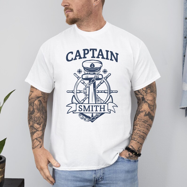 May include: White t-shirt with a navy blue nautical design. The design features the word "CAPTAIN" above an anchor, ship's wheel, and the name "SMITH" in a banner. The shirt has short sleeves and a crew neck.