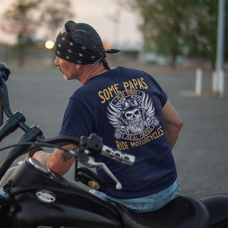 Puede incluir: Una camiseta azul marino con un gr&aacute;fico dorado de una calavera con casco y sosteniendo una llave inglesa. El texto en la camiseta dice "Some Papas Play Bingo Real Papas Ride Motorcycles".