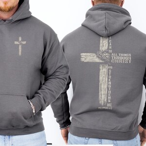 K&ouml;nnte beinhalten: Grauer Kapuzenpullover mit Fronttasche und Kordelzugkapuze. Auf der Vorderseite befindet sich ein kleines, helles Kreuz. Auf der R&uuml;ckseite ist ein gr&ouml;&szlig;eres Kreuzdesign mit dem Text "I CAN DO ALL THINGS THROUGH CHRIST WHO STRENGTHENS ME" und "PHILIPPIANS 4:13" abgebildet.