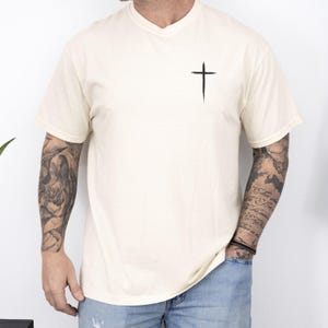 May include: Cream-colored V-neck t-shirt with a black cross graphic on the chest. The shirt is made of a soft material and is suitable for casual wear. The model is wearing blue jeans.