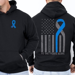 May include: Black hooded sweatshirt with a blue ribbon graphic on the front and back. The back features a distressed American flag design with the text "T1D Awareness" and "NO ONE FIGHTS ALONE".