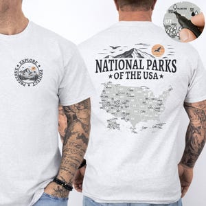Peut inclure: T-shirt gris clair avec un motif sur le devant : "EXPLORE PRESERVE PROTECT" autour d'une montagne. Le dos affiche "NATIONAL PARKS OF THE USA" au-dessus d'une carte des États-Unis avec les parcs.