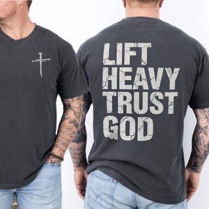 Pepper Comfort Colors style Christian gym shirt shown on a tattooed man, small distressed cross on the front chest and large cracked white text on the back reading “LIFT HEAVY TRUST GOD,” casual short sleeve tee with a relaxed fit.