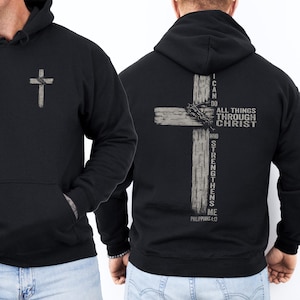 K&ouml;nnte beinhalten: Schwarzer Kapuzenpullover mit einer Vordertasche und einer grauen Kreuzgrafik auf der Vorderseite. Auf der R&uuml;ckseite befindet sich ein gr&ouml;&szlig;eres graues Kreuzdesign mit einer Dornenkrone und dem Text "I CAN DO ALL THINGS THROUGH CHRIST WHO STRENGTHENS ME" aus Philipper 4:13.