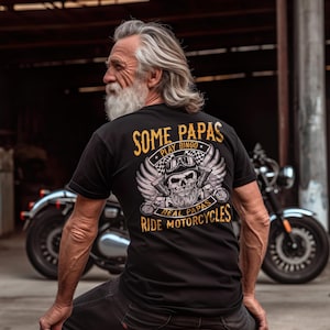 Puede incluir: Camiseta negra con un gr&aacute;fico dorado de una calavera con barba y alas. El texto "Some Papas Play Bingo Real Papas Ride Motorcycles" est&aacute; impreso en dorado en la camiseta.