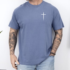 May include: A short-sleeved, blue t-shirt with a white cross graphic on the upper left chest. The shirt is made of a soft material and has a relaxed fit. The model is wearing blue jeans.