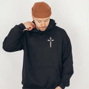 K&ouml;nnte beinhalten: Schwarzer Kapuzenpullover mit einem wei&szlig;en Kreuz-Grafik auf der Vorderseite. Der Pullover hat eine K&auml;ngurutasche und eine Kapuze mit Kordelzug. Die Person im Bild tr&auml;gt eine braune M&uuml;tze.