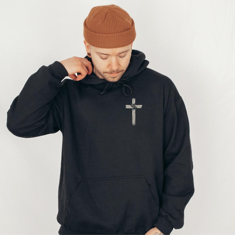 K&ouml;nnte beinhalten: Schwarzer Kapuzenpullover mit einem wei&szlig;en Kreuz-Grafik auf der Vorderseite. Der Pullover hat eine K&auml;ngurutasche und eine Kapuze mit Kordelzug. Die Person im Bild tr&auml;gt eine braune M&uuml;tze.