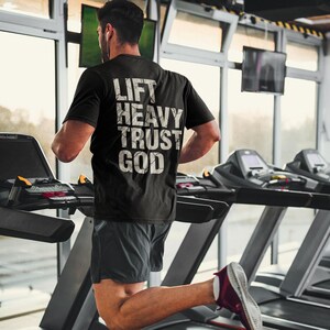 May include: Black t-shirt with the text "LIFT HEAVY TRUST GOD" in white, worn by a person running on a treadmill. The person is wearing gray shorts and red and white sneakers. The background shows a gym setting with treadmills and windows.