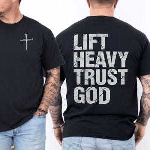 May include: Black t-shirt with a white cross graphic on the front and the text "LIFT HEAVY TRUST GOD" on the back. The text has a distressed, cracked appearance. The shirt is a crew neck style.