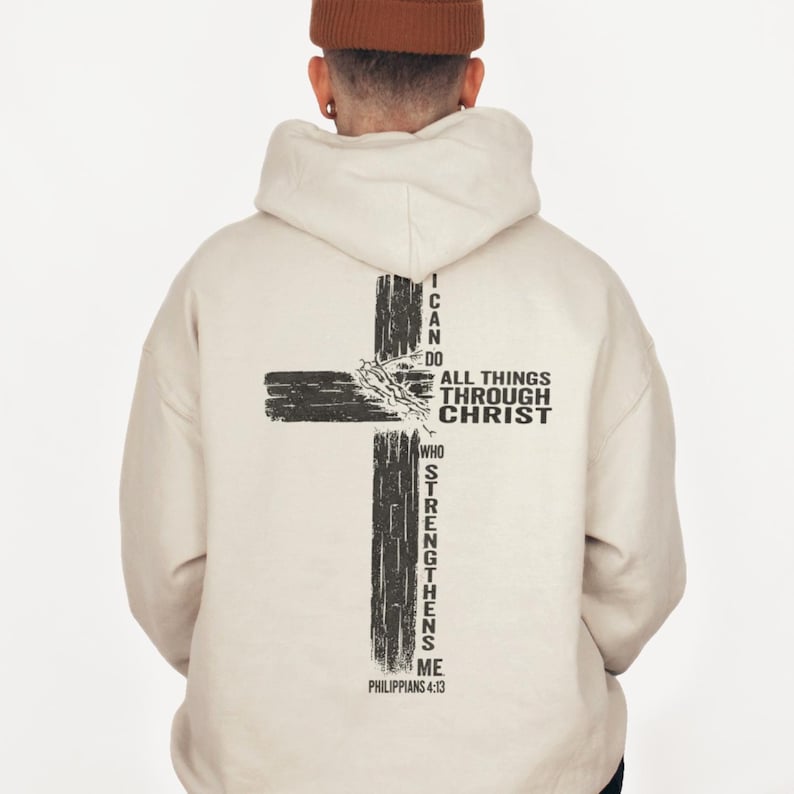 Man wearing a beige Christian hoodie with a large distressed black cross on the back, featuring praying hands and the Bible verse “I can do all things through Christ who strengthens me – Philippians 4:13”