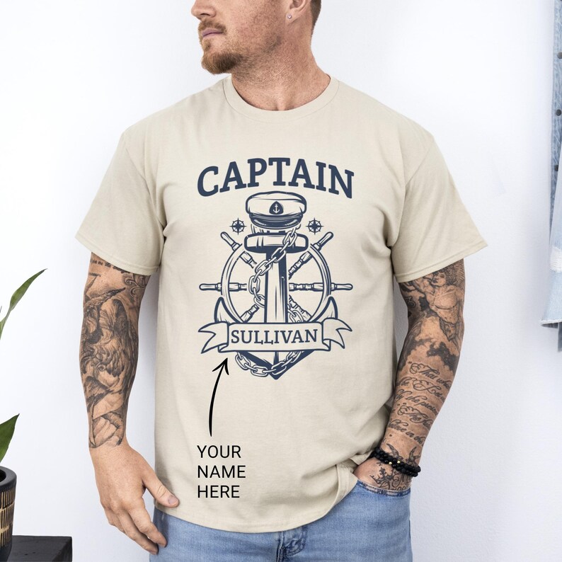 Sand Colored cotton t-shirt with bold navy graphic featuring a captain’s hat, anchor, ship wheel, and personalized name banner reading “SULLIVAN,” worn by a tattooed man; custom captain shirt ideal for boat owners or as a Father’s Day gift.