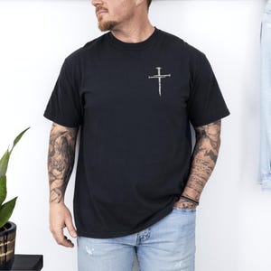 May include: Black short-sleeved t-shirt with a small, silver-colored cross design on the upper left chest. The cross is made to look like it is made of nails. The shirt is a solid black color.