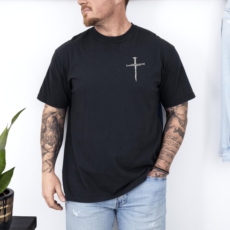 May include: Black short-sleeved t-shirt with a small, silver-colored cross design on the upper left chest. The cross is made to look like it is made of nails. The shirt is a solid black color.