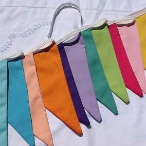 May include: A colourful fabric bunting with flags in shades of teal, blue, peach, orange, purple, lavender, green, lime green, pink, and yellow.