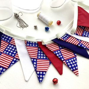 May include: A fabric bunting banner featuring triangular flags in solid red, solid white, solid navy blue, and a pattern of the American flag with stars and stripes. The banner is accompanied by a silver tassel, a spool of white thread, and small red and blue jingle bells.