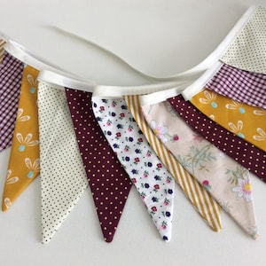 May include: A fabric bunting banner featuring a series of triangular flags in various patterns including mustard yellow floral, burgundy polka dot, white with small blue and red flowers, cream with black dots, and gold and white diagonal stripes.