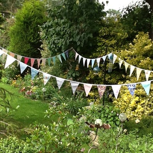 May include: A string of colourful fabric bunting flags strung between trees in a garden setting. The flags are made of different patterns and colours, including pink, blue, green, and white.