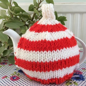 May include: A white ceramic teapot is covered with a red and white striped knitted tea cozy. The cozy has a pom-pom on top. The teapot is on a floral patterned tablecloth, with green foliage in the background.